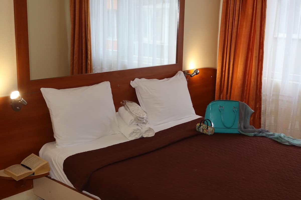 A hotel room with a bed, pillows, a nightstand, a lamp, and a window with curtains.