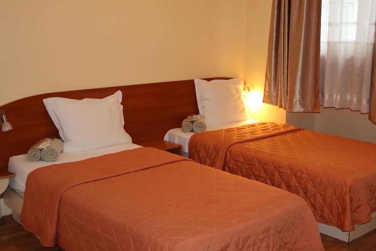 A hotel room with two single beds, each with a wooden headboard, white linens, and an orange quilted bedspread. There are two bedside tables with lamps, and the room has a warm, neutral color scheme.