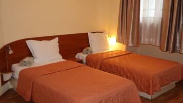 A hotel room with two single beds, each with a wooden headboard, white linens, and an orange quilted bedspread. There are two bedside tables with lamps, and the room has a warm, neutral color scheme.