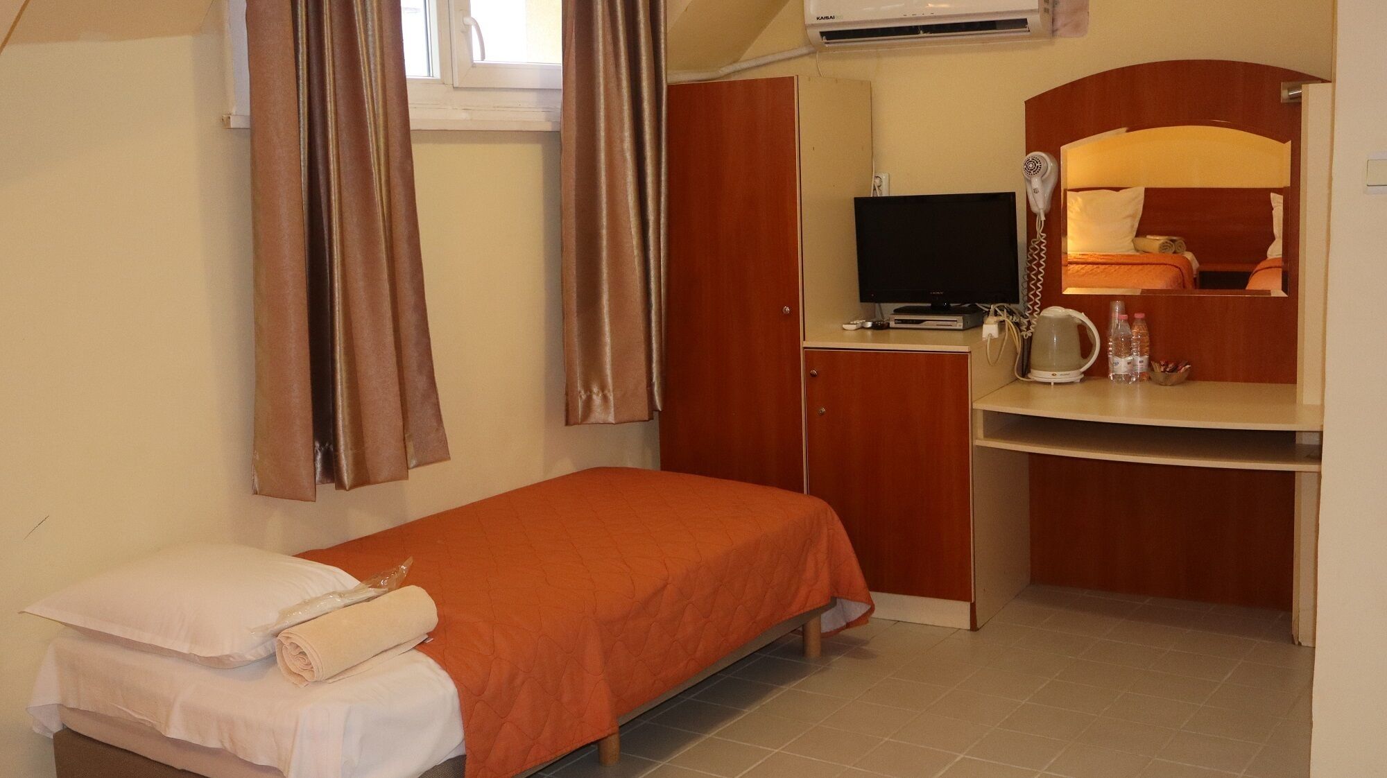 Una habitación pequeña con cama individual, un armario de madera, un escritorio con computadora y un espejo, y una ventana con cortinas.