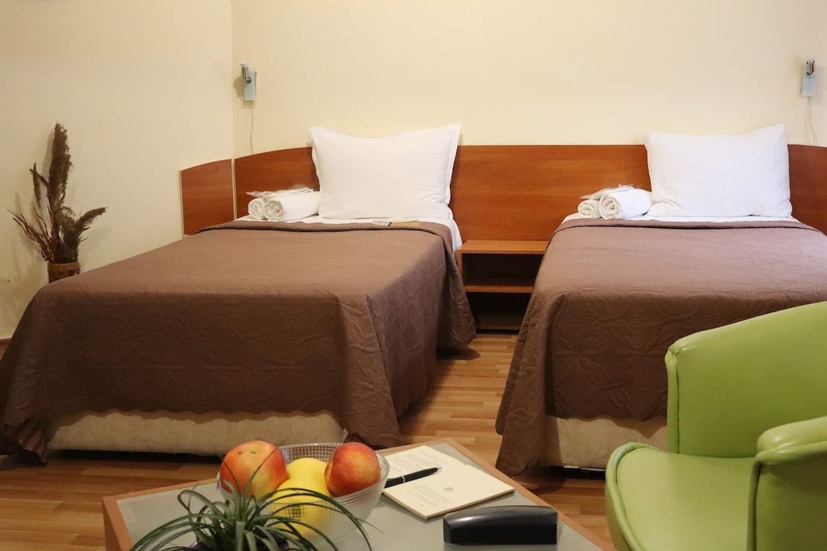 A hotel room with two beds, a green chair, a table with fruits and a pen, and a plant.