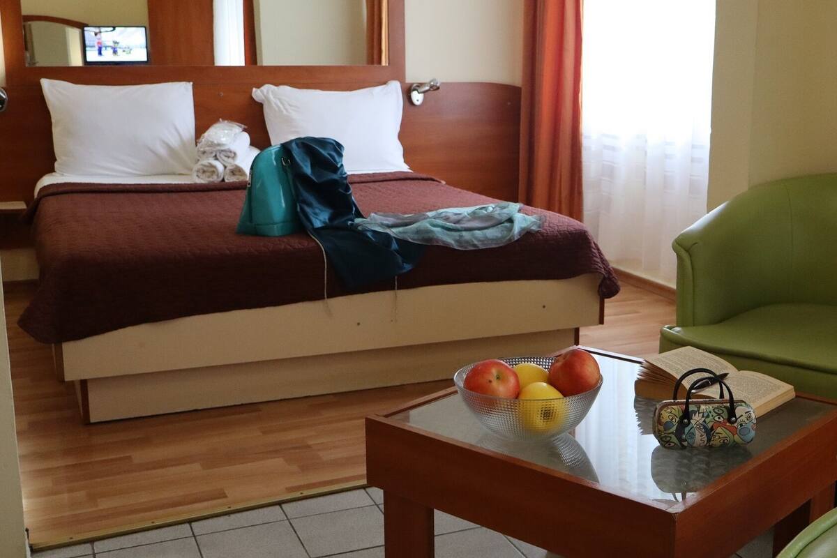 A hotel room with a bed, a green armchair, a small table with a fruit bowl and a book, and a television on the wall.