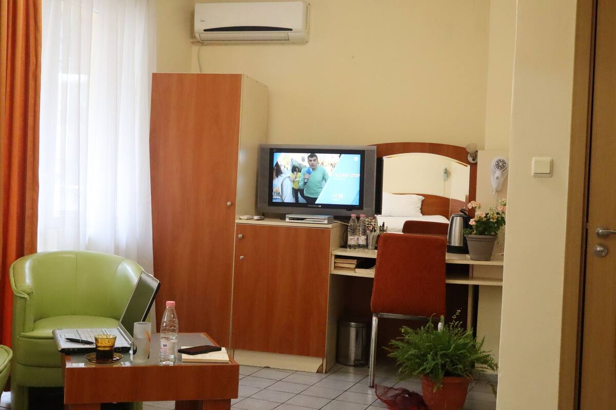 A hotel room with a television, a desk with a laptop, a green chair, a wooden wardrobe, and a bed with a headboard.