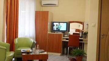 Una habitación de hotel con televisión, un escritorio con computadora portátil, una silla verde, un armario de madera y una cama con cabecera.