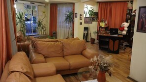 A living room with a brown sofa, a glass coffee table, and a potted plant. There is a desk with a computer and a teddy bear. The room has a wooden floor and orange curtains. - Palitra Family Hotel (Varna)