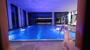 A poolside view with a person relaxing on a lounge chair, a pool with blue lighting, and a view of the night sky. - Hotel Nova (Gaschurn)