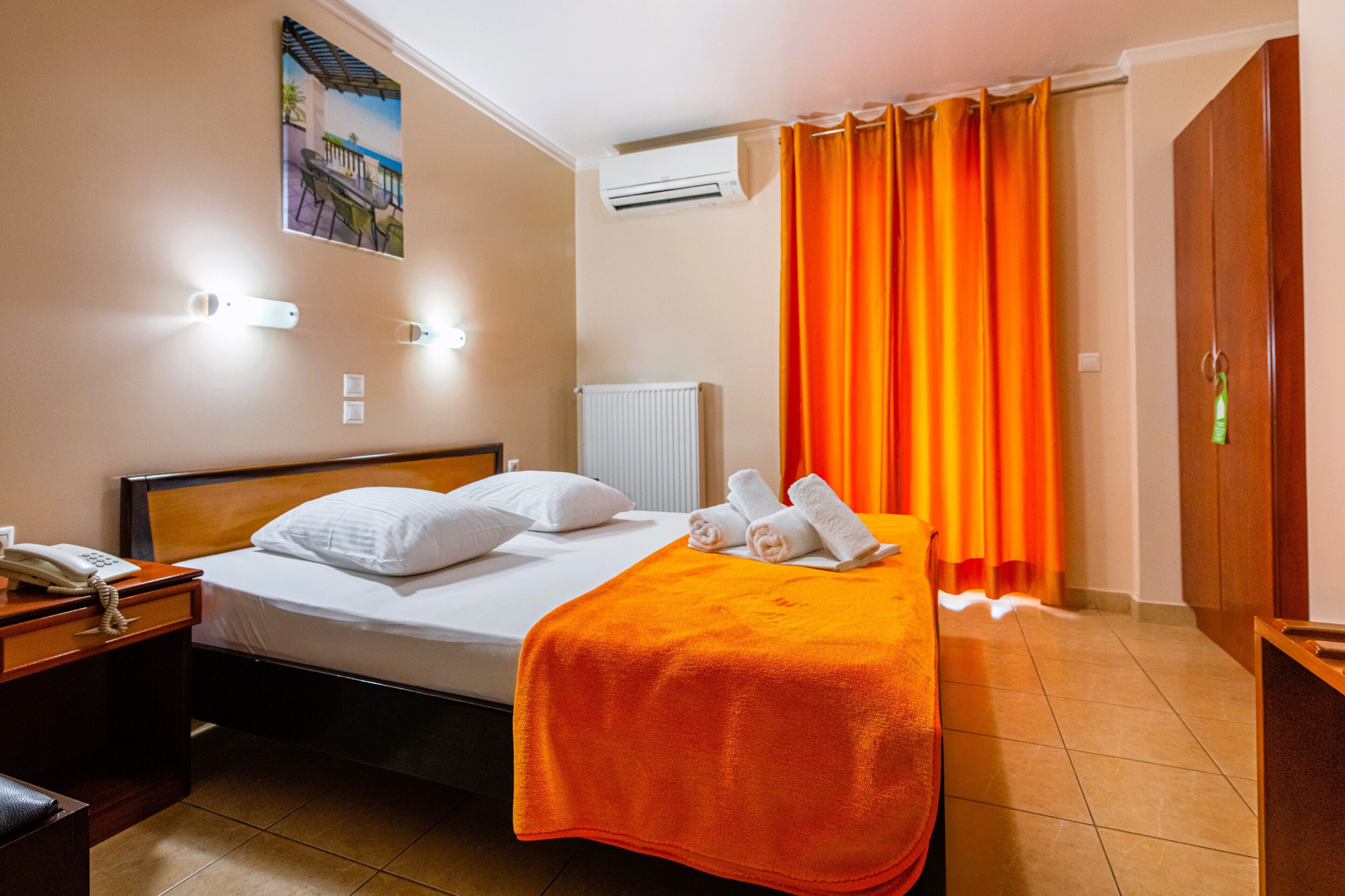 A hotel room with a bed, a nightstand with a telephone, an air conditioning unit, and a painting on the wall.