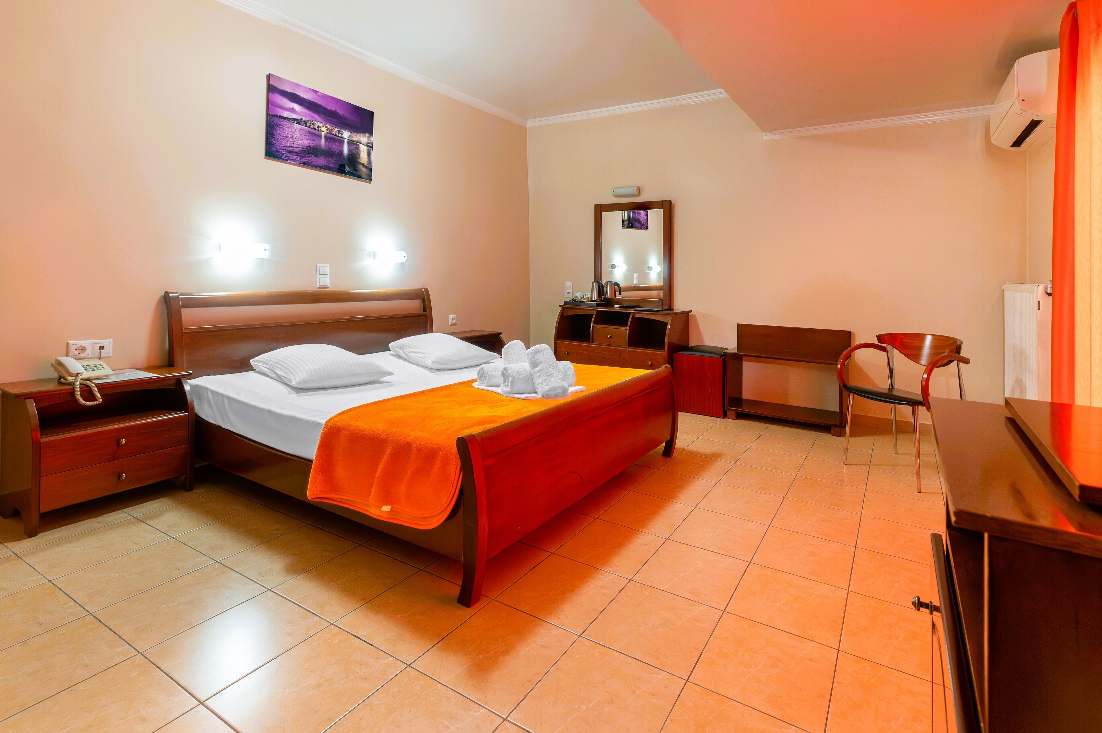 Habitación de hotel con una cama, una mesita de noche con teléfono, una silla, un escritorio, un espejo y un aire acondicionado de pared.