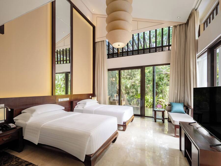 A hotel room with two beds, a TV, a desk, and a balcony with a view of greenery.
