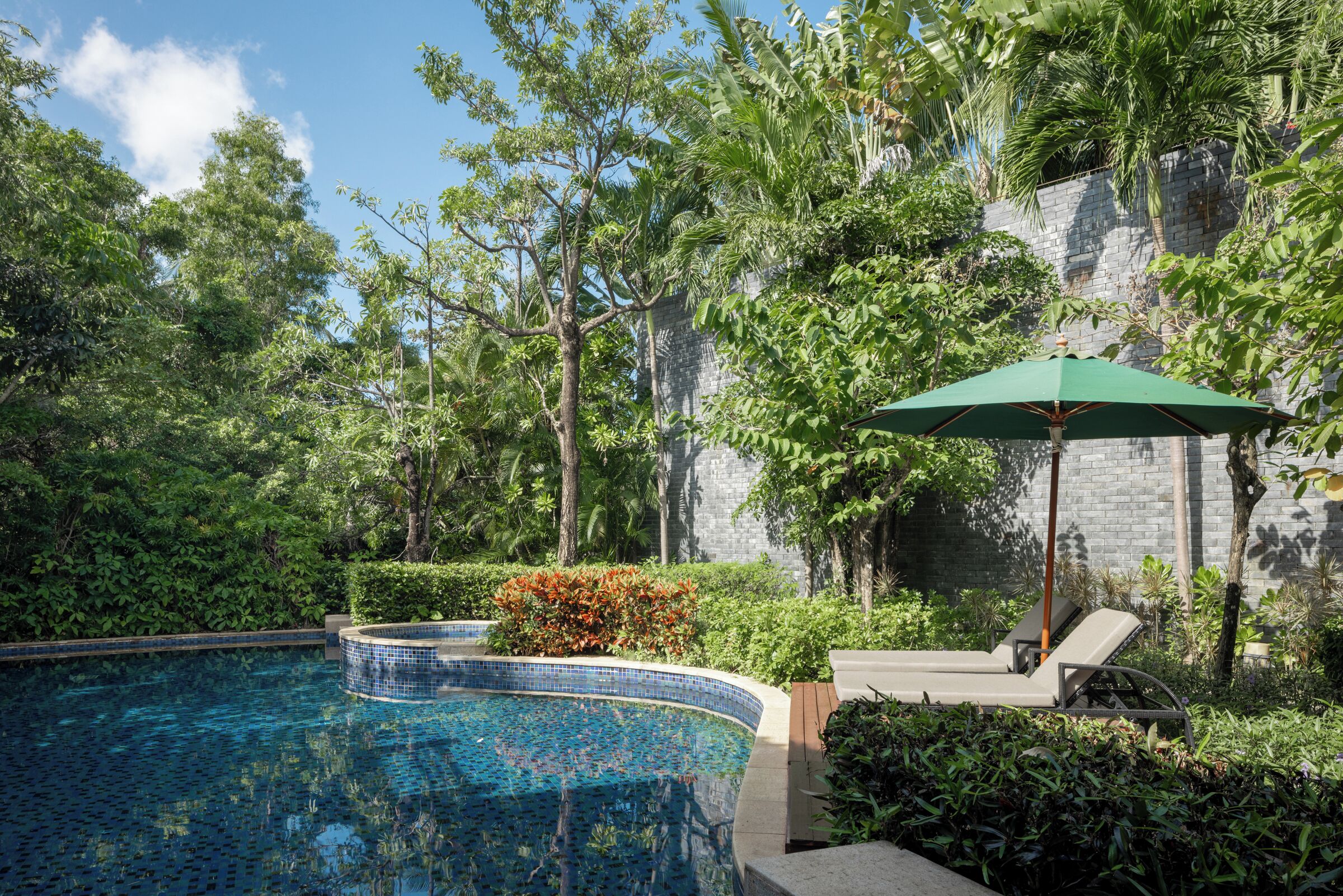 A swimming pool with a tiled edge, surrounded by lush greenery and a stone wall.