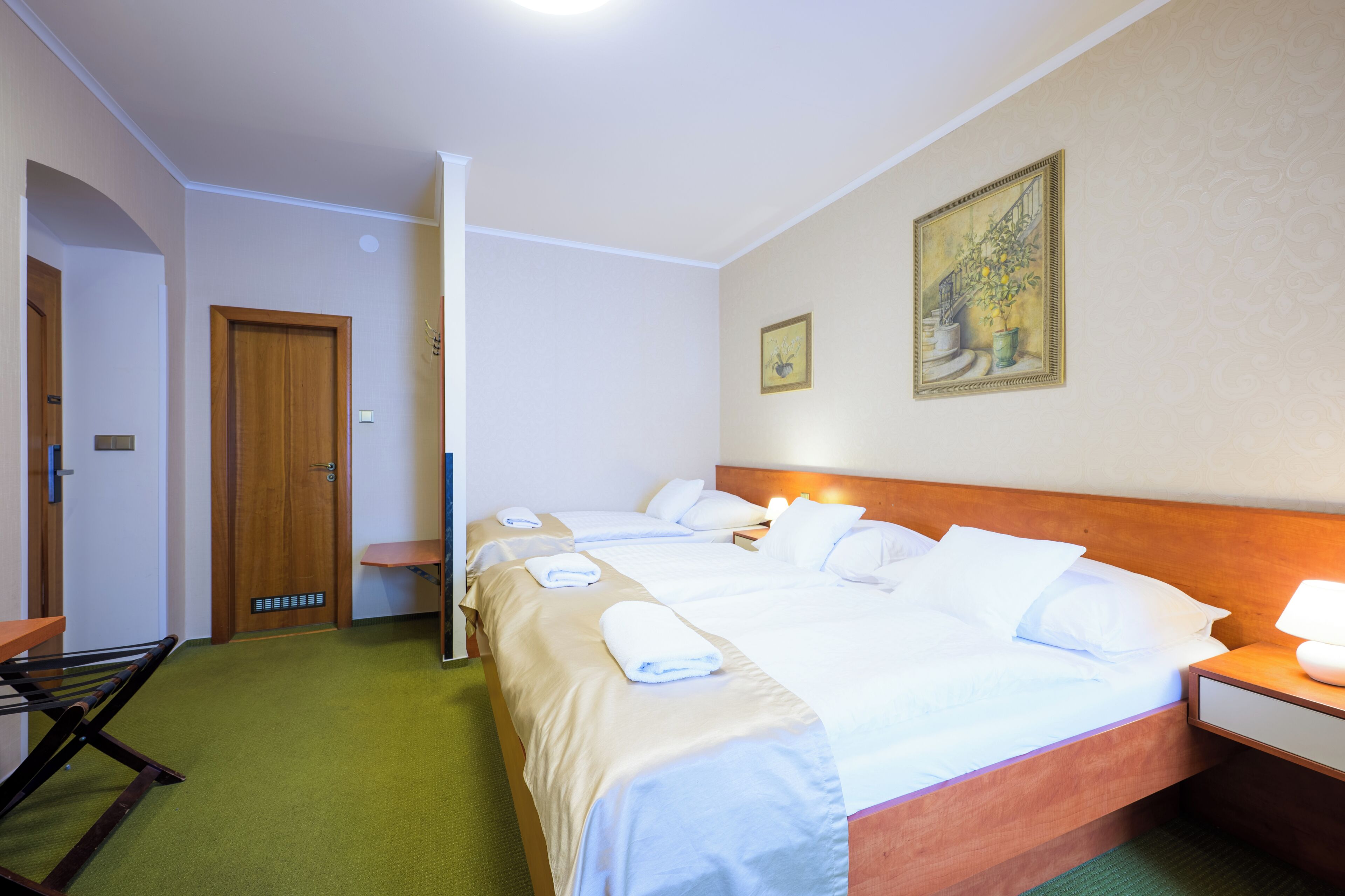 Ein Hotelzimmer mit zwei Betten, einem hölzernen Kopfteil, einem Nachttisch mit Lampe, einem Bild an der Wand und einem grünen Teppich.