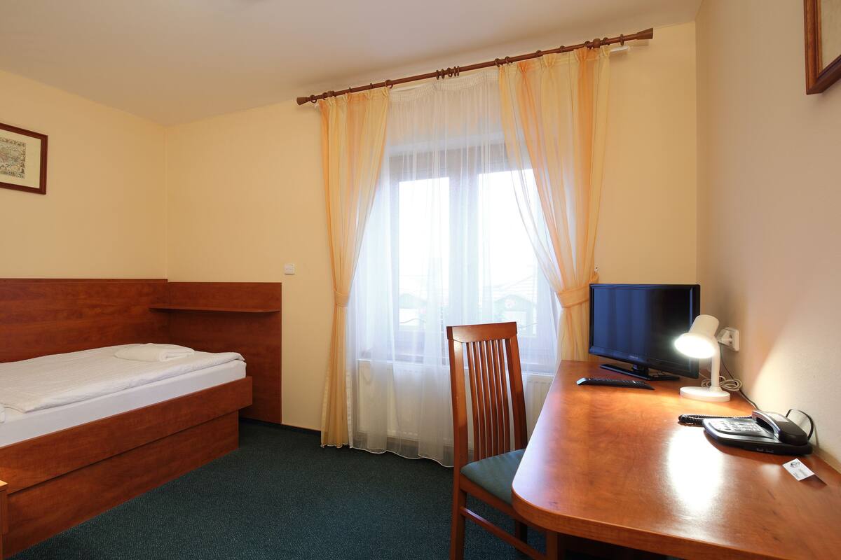 A hotel room with a single bed, a desk with a computer and phone, a chair, and a window with sheer curtains.