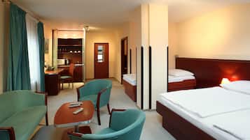 A hotel room with a bed, two green armchairs, a wooden table, a desk, and a kitchenette.