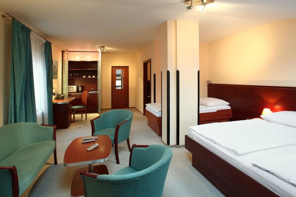 A hotel room with a bed, two green armchairs, a wooden table, a desk, and a kitchenette.