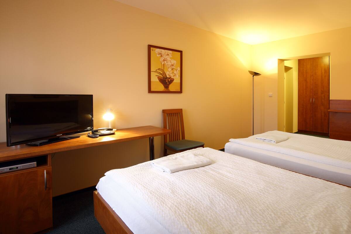 A hotel room with a TV, desk, chair, two beds, and artwork on the walls.