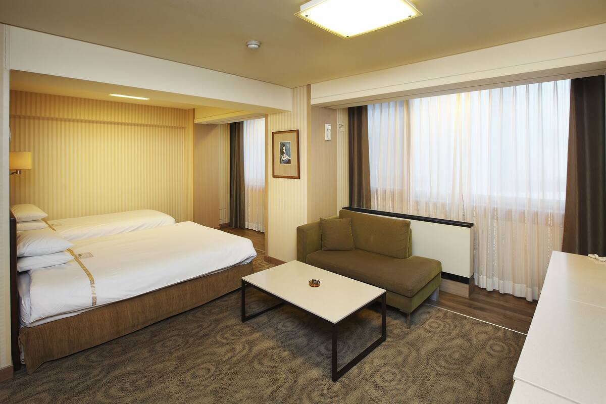 A hotel room with a double bed, a sofa, a small table, and a large window with curtains.