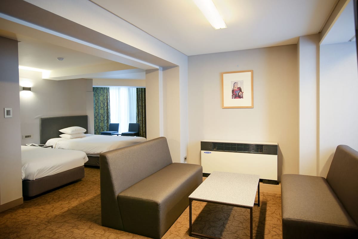 A hotel room with two single beds, a sofa, a small table, and a wall-mounted air conditioner.