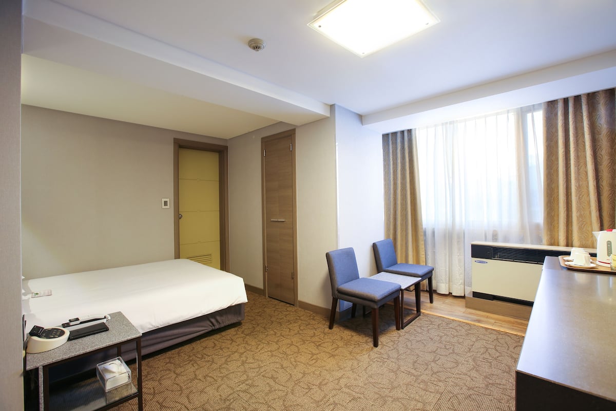 A hotel room with a bed, two chairs, a desk with a telephone, and an air conditioning unit.