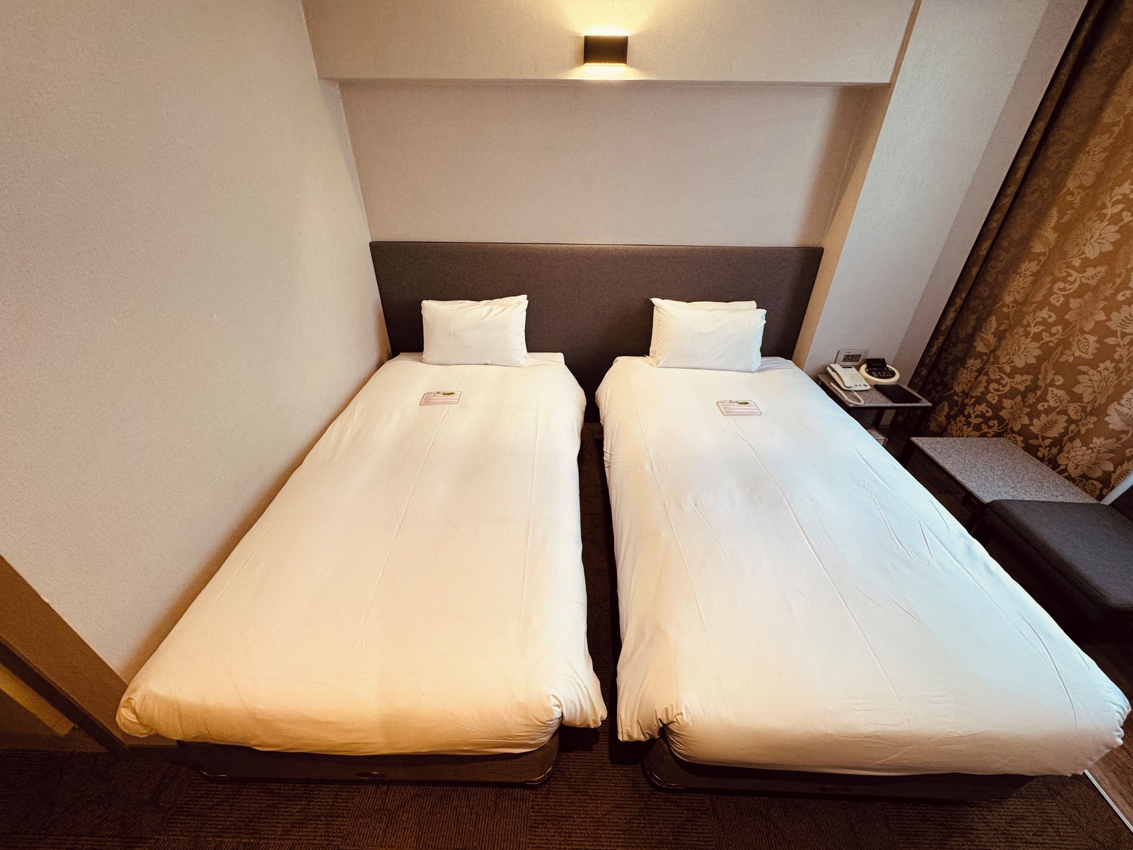 A twin room with two single beds, a bedside table with a lamp, and a small desk with a phone and remote controls.