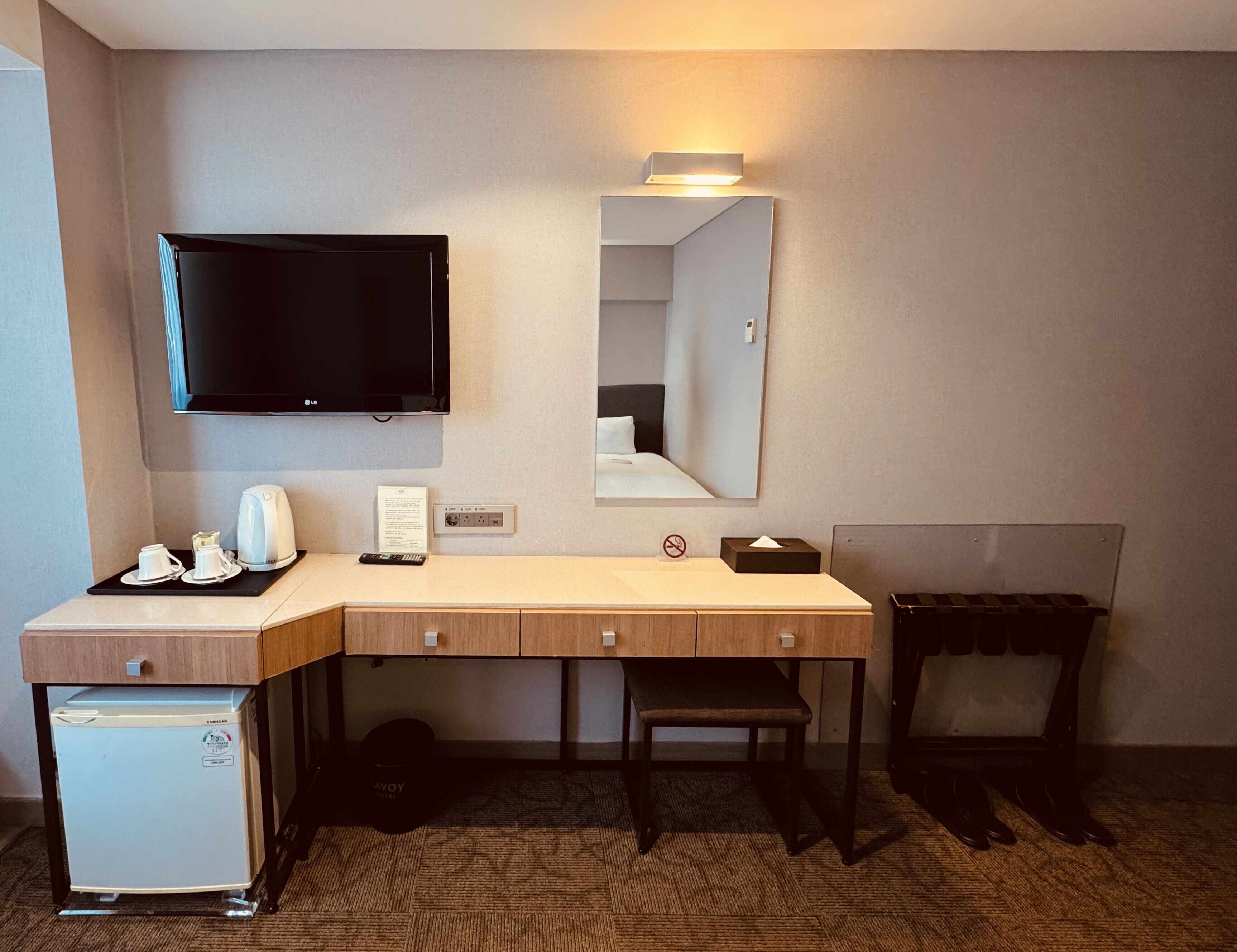 Habitación de hotel con escritorio, televisión, espejo, refrigerador pequeño y cubo de basura.