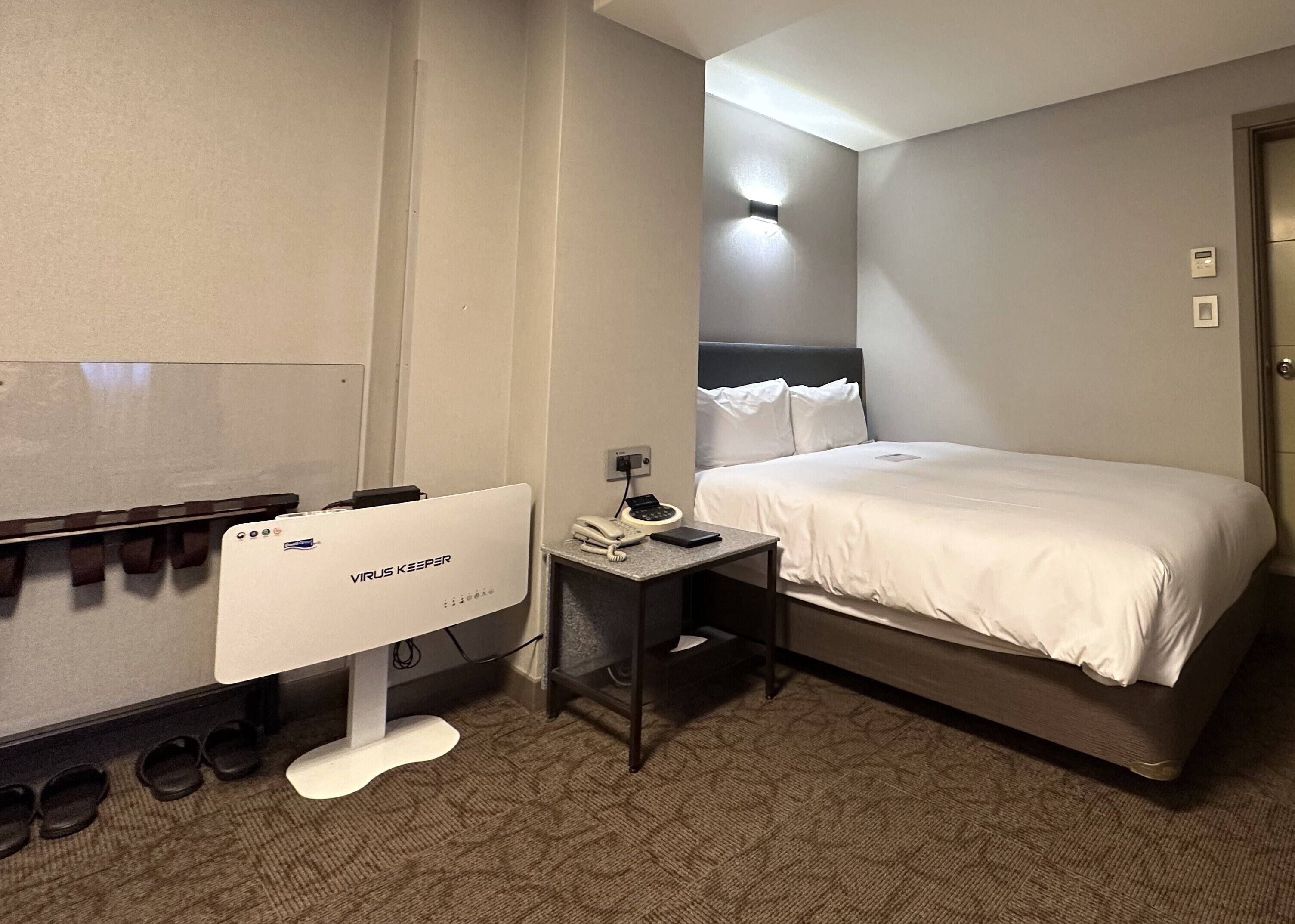 A hotel room with a bed, a desk with a phone, a computer, and a shoe rack.