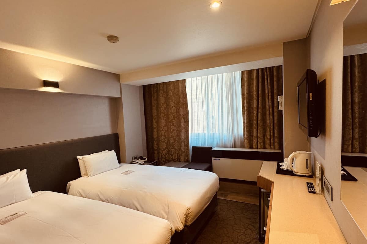 A hotel room with two beds, a desk, a television, and a window with curtains.