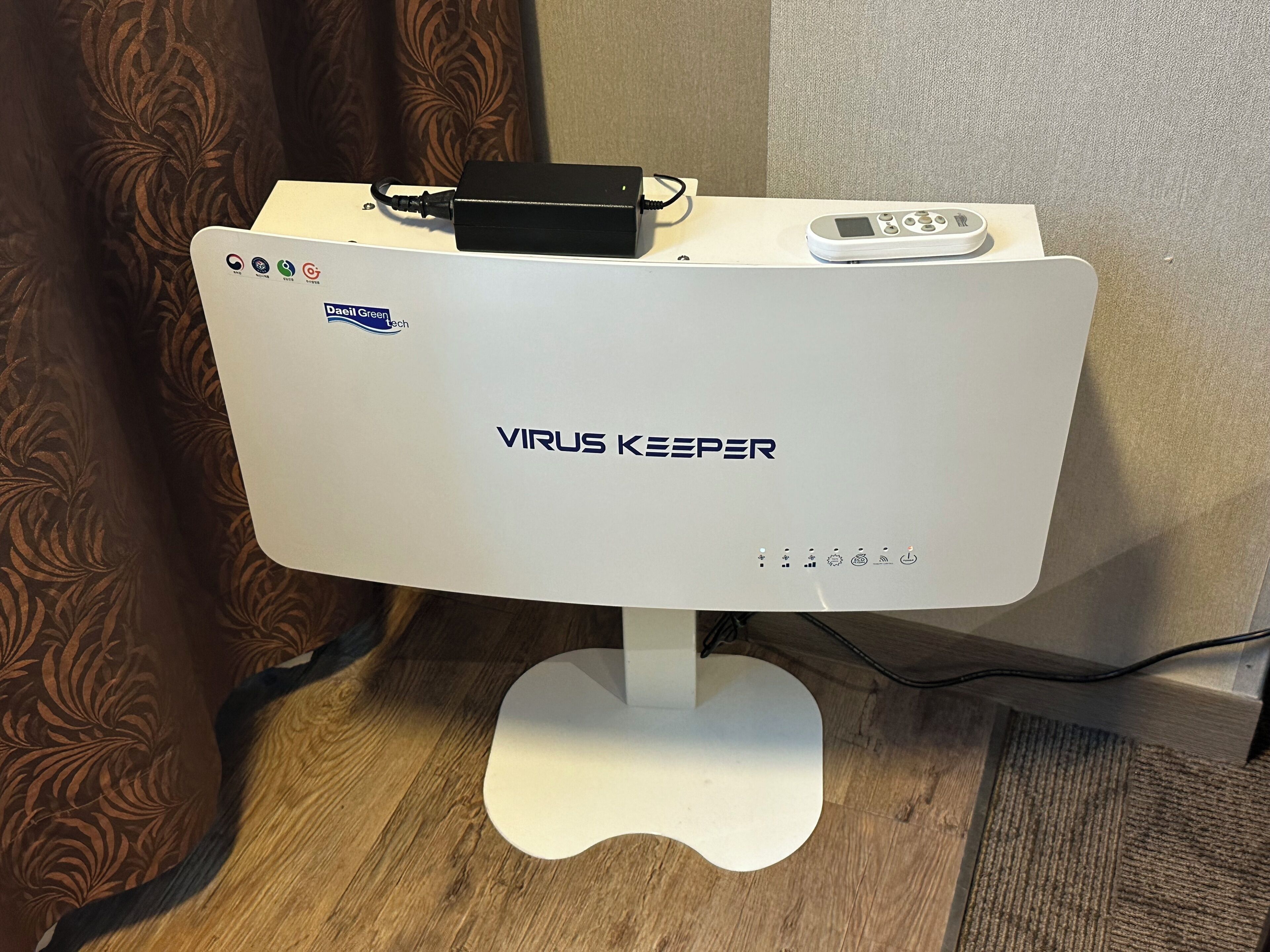 Un dispositivo blanco con la etiqueta "VIRUS KEEPER" sobre una base blanca, con un control remoto y un adaptador de corriente negro encima.