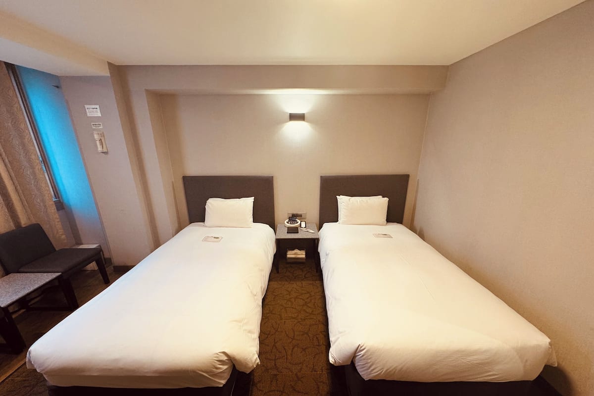 A hotel room with two single beds, a small desk, and a chair.