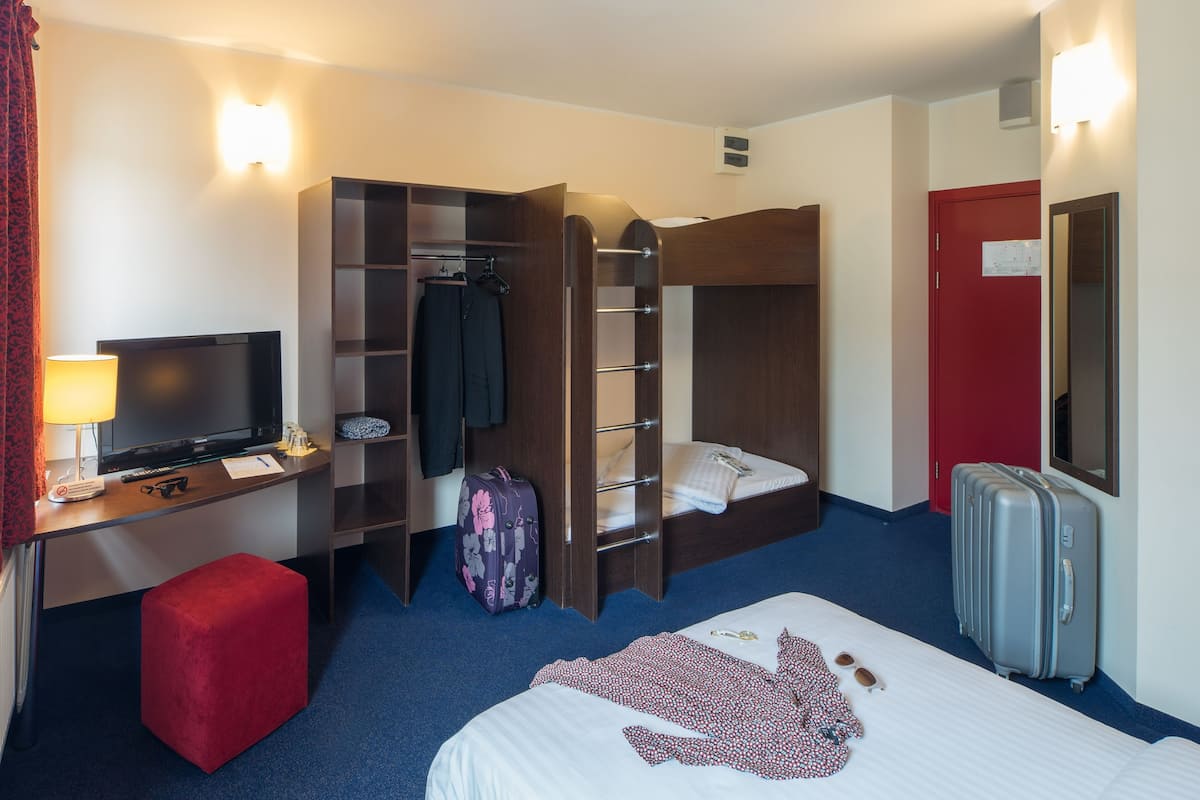 A hotel room with a bunk bed, a desk with a TV, a red ottoman, and a suitcase.