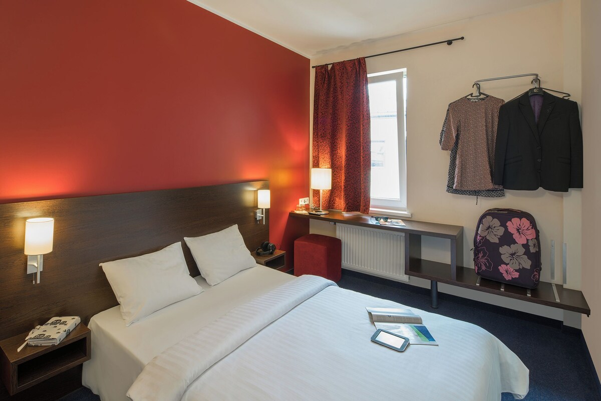 A hotel room with a bed, bedside lamps, a desk with a lamp, a suitcase, and a coat rack with jackets.