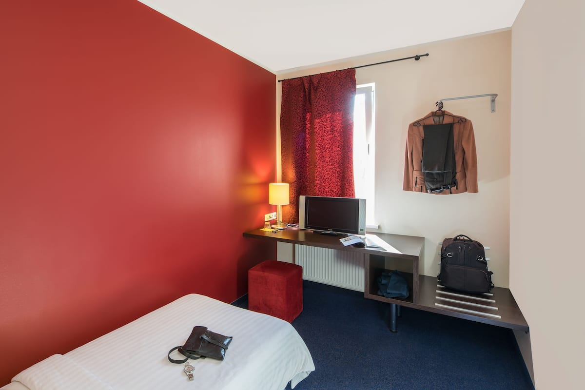 A hotel room with a bed, a desk with a TV, a red wall, a red curtain, and a coat rack with a jacket and a backpack.