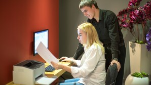 Una persona sentada en un escritorio con una computadora, otra persona de pie a su lado, e una impresora sobre el escritorio.