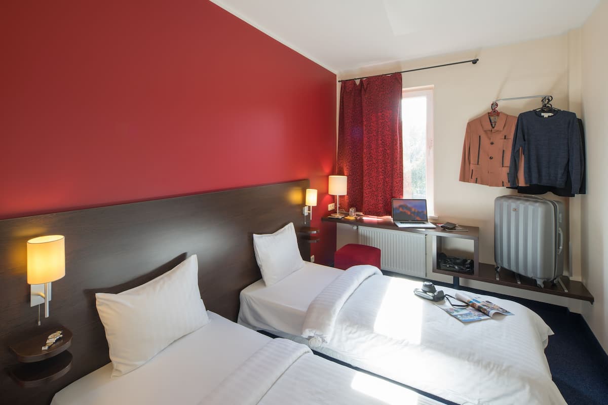A hotel room with two beds, a desk with a computer, a red curtain, and a coat rack with two jackets.