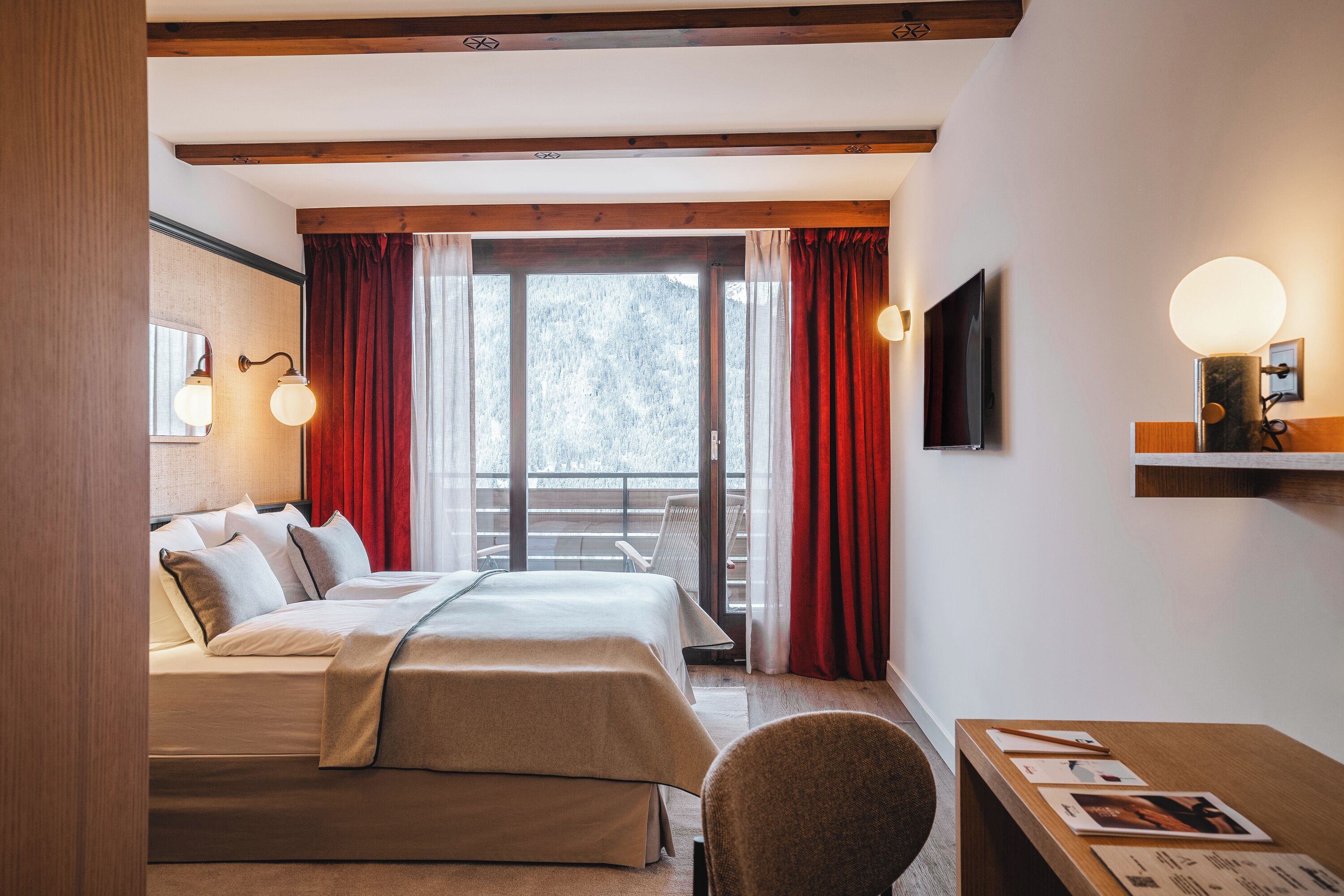 A hotel room with a bed, a desk, a chair, a TV, and a balcony with red curtains.