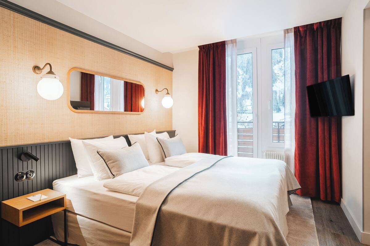 Une chambre d’hôtel avec un grand lit, une table de chevet, un miroir, des appliques murales, un téléviseur à écran plat et une fenêtre avec des rideaux rouges.