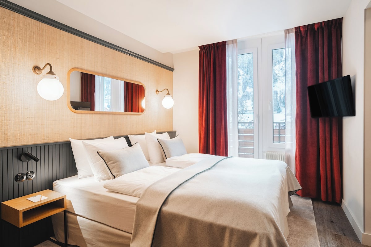 Ein Hotelzimmer mit einem Bett, Nachttischen, einem Spiegel, Wandleuchten, einem Fernseher und einem Fenster mit Vorhängen.