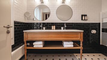 Un baño con pared de azulejos blancos, suelo a cuadros blanco y negro, tocador de madera con dos lavabos y un espejo sobre el tocador.