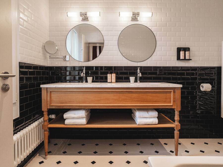 Un baño con pared de azulejos blancos, suelo a cuadros blanco y negro, tocador de madera con dos lavabos y un espejo sobre el tocador.
