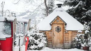 Une cabane en bois dont le toit est recouvert de neige, un véhicule rouge avec des skis garé à proximité, et des arbres chargés de neige en arrière-plan.