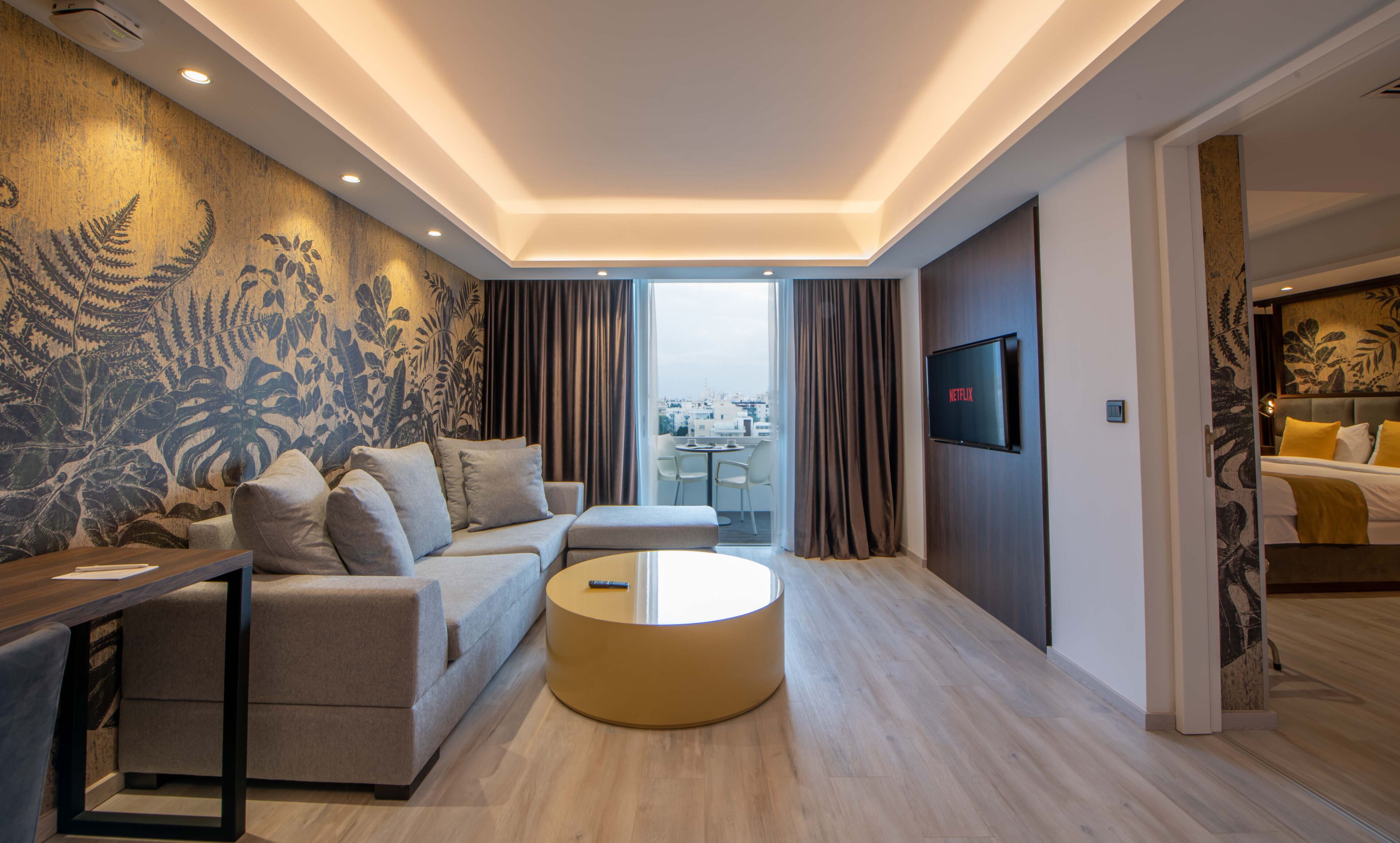 A modern living room with a sofa, a round coffee table, a TV mounted on the wall, and a balcony with a view of the city.