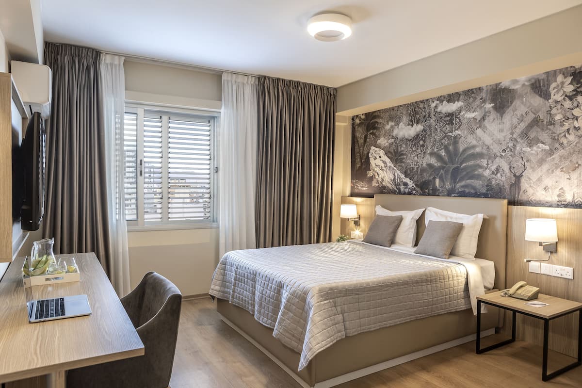 A hotel room with a bed, bedside lamps, a desk with a laptop, and a large abstract painting on the wall.