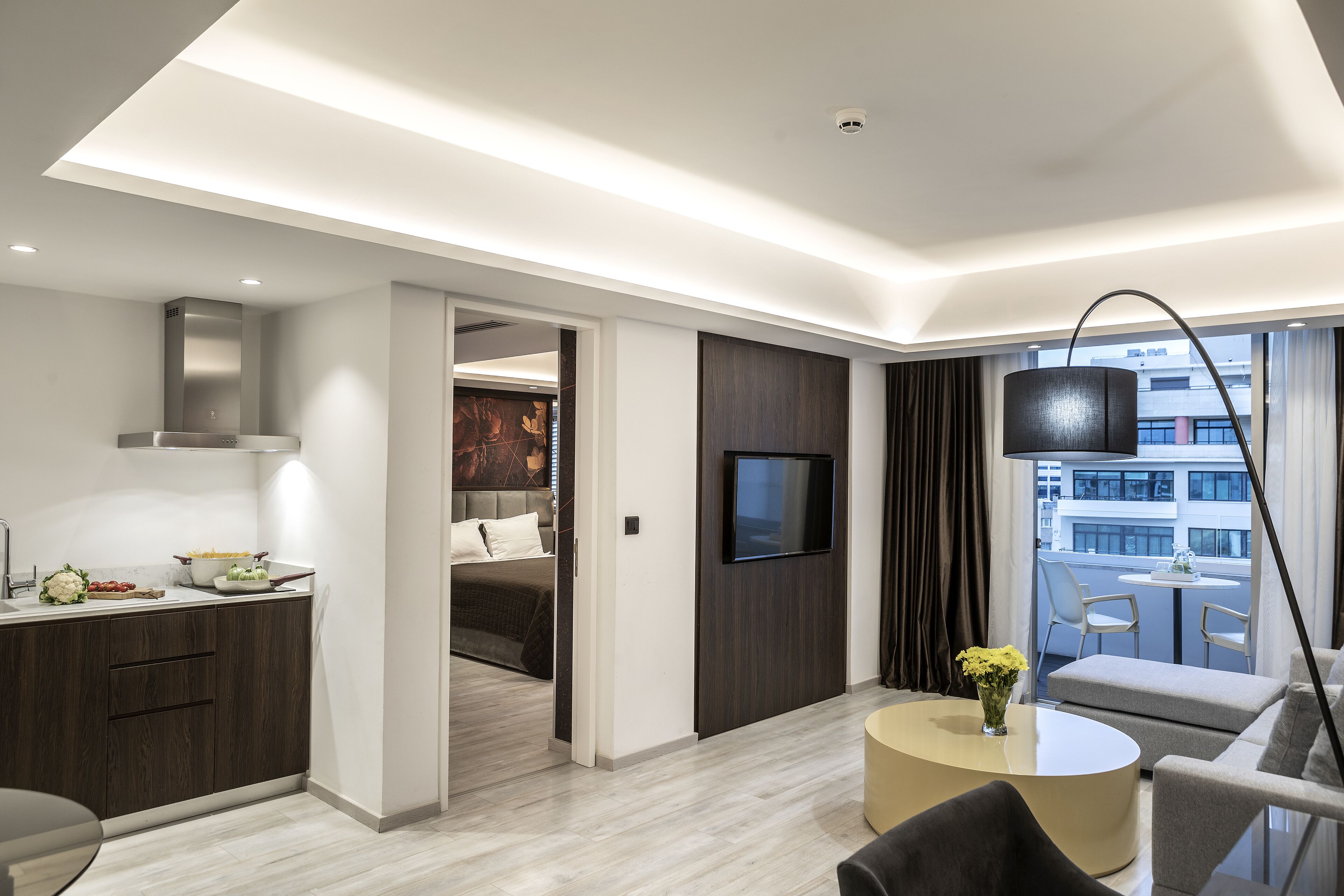 A modern hotel room with a kitchen area, a dining table, and a living area with a sofa and a TV.