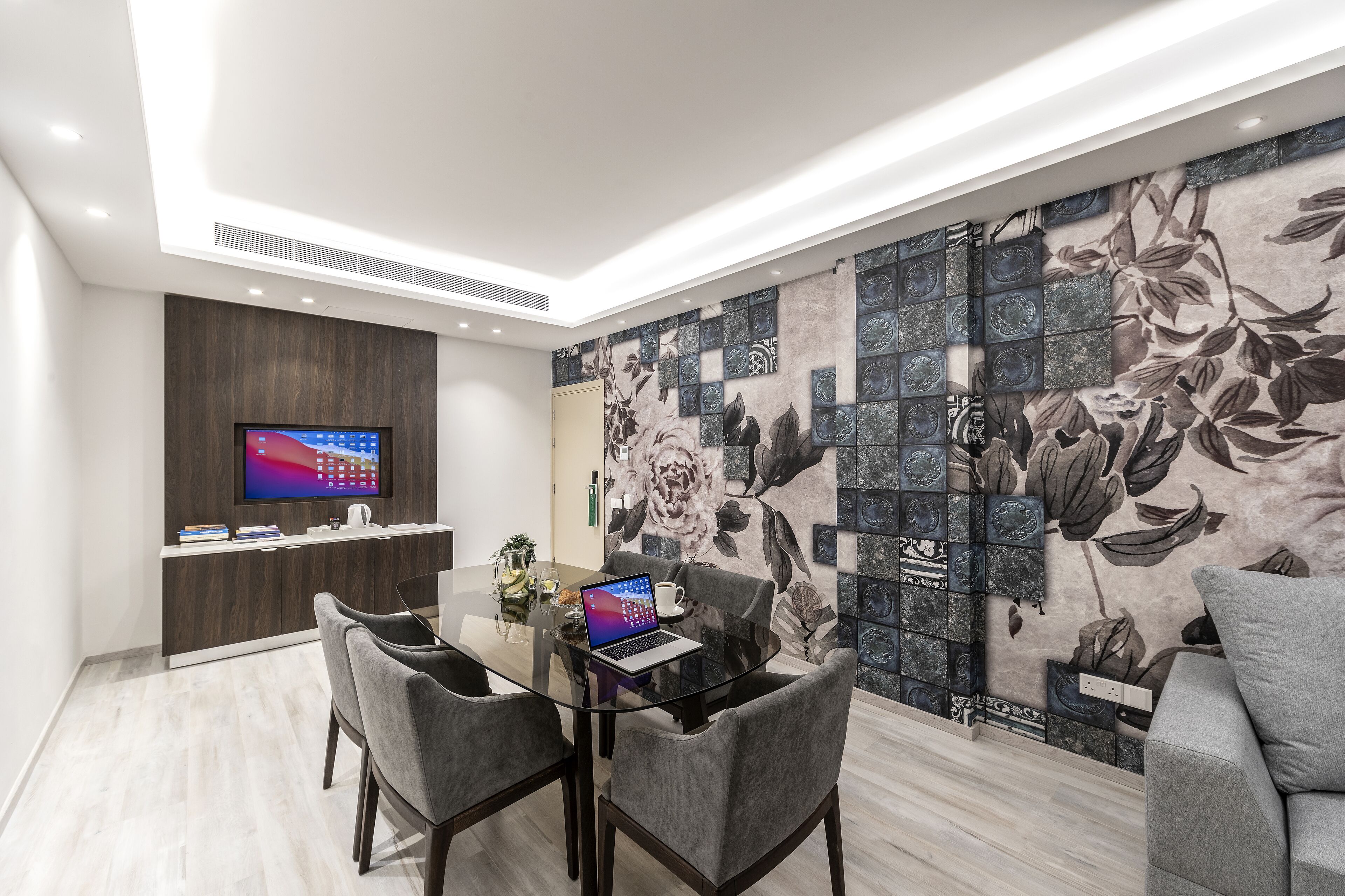 Quarto moderno com mesa de jantar de vidro, cadeiras estofadas cinzentas, televisão de tela plana e parede decorativa com azulejos florais.