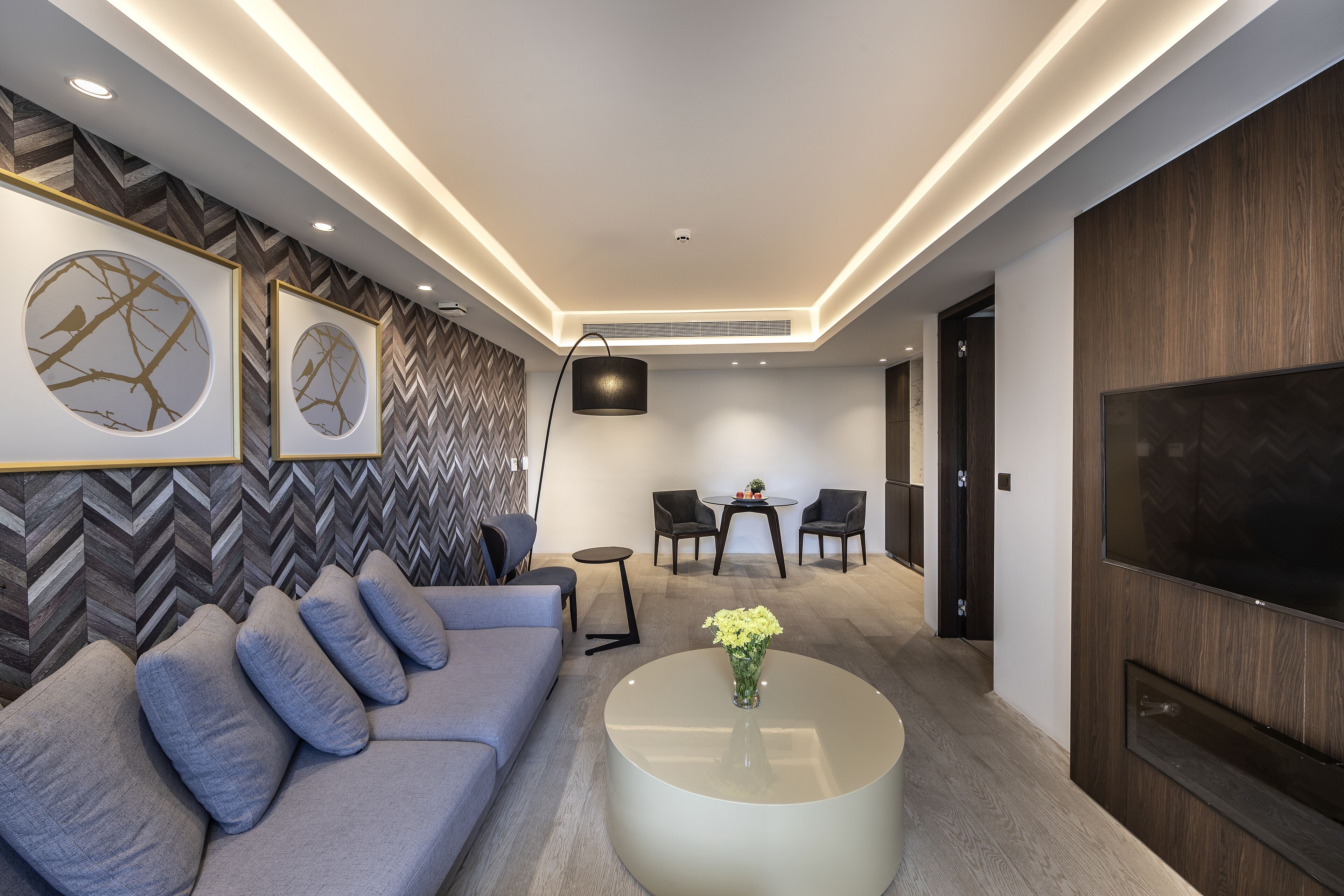 Un salon moderne avec un canapé gris, une table basse ronde ornée de fleurs, un téléviseur et un mur décoré d’un motif en chevrons.