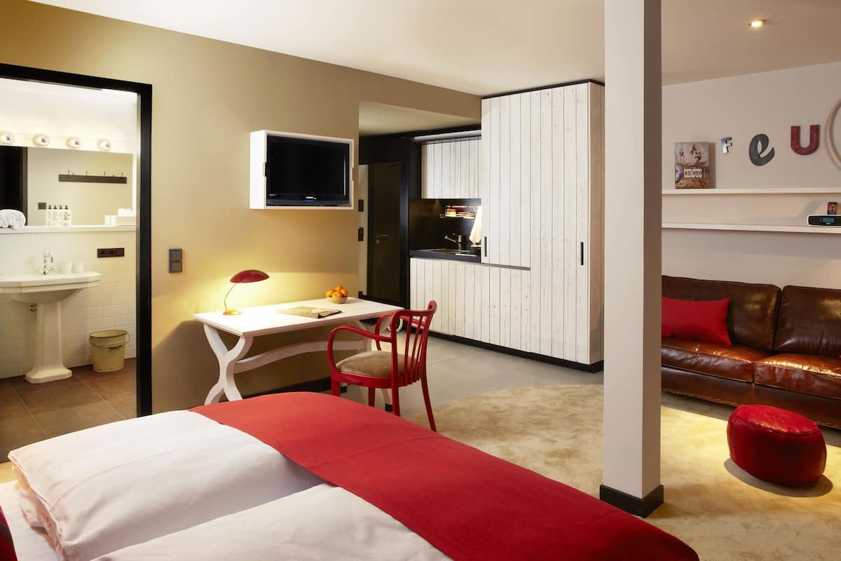 A modern hotel room with a bed, a desk, a red chair, a red ottoman, and a kitchenette.