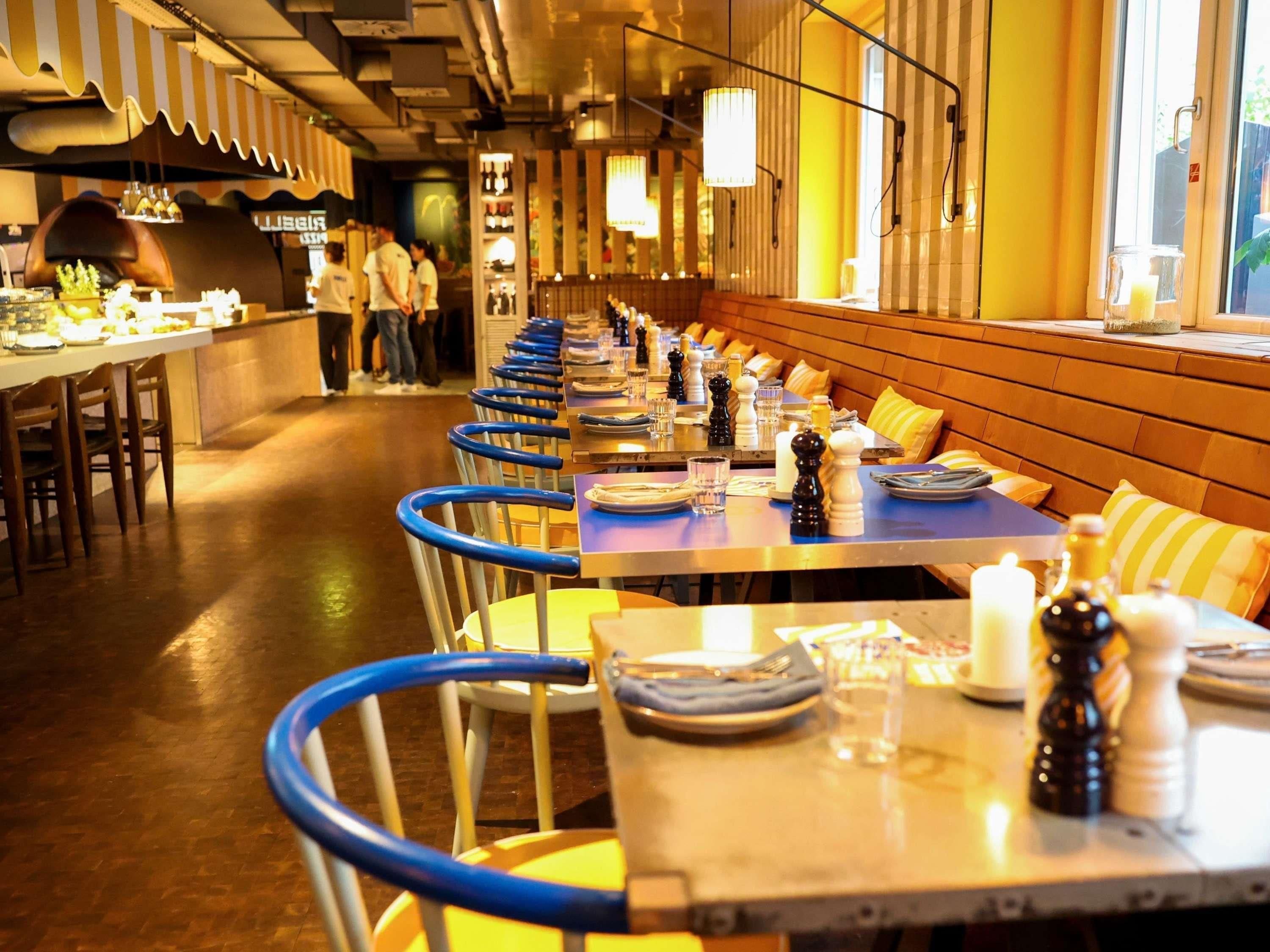 Um restaurante com mesas compridas, cadeiras azuis e uma fileira de cabines com almofadas.