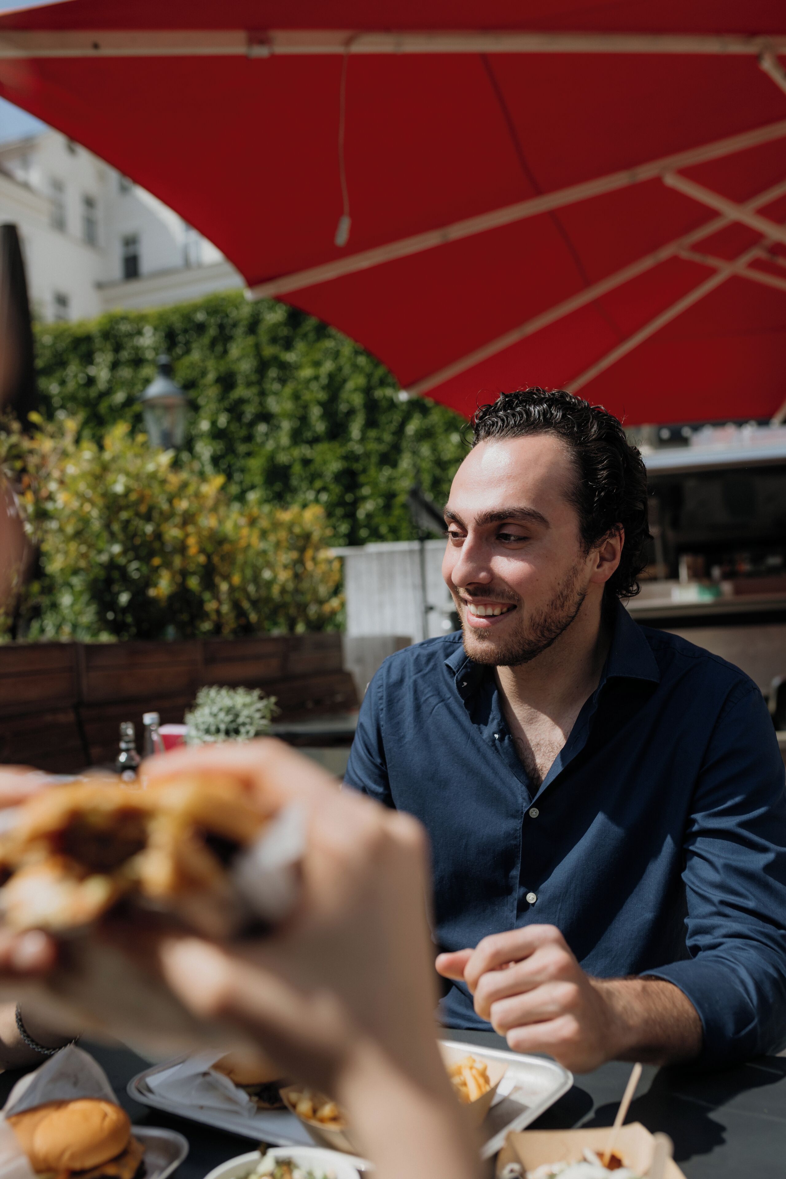 Une personne est assise à une table à l’extérieur, devant un repas, sous un parasol rouge.
