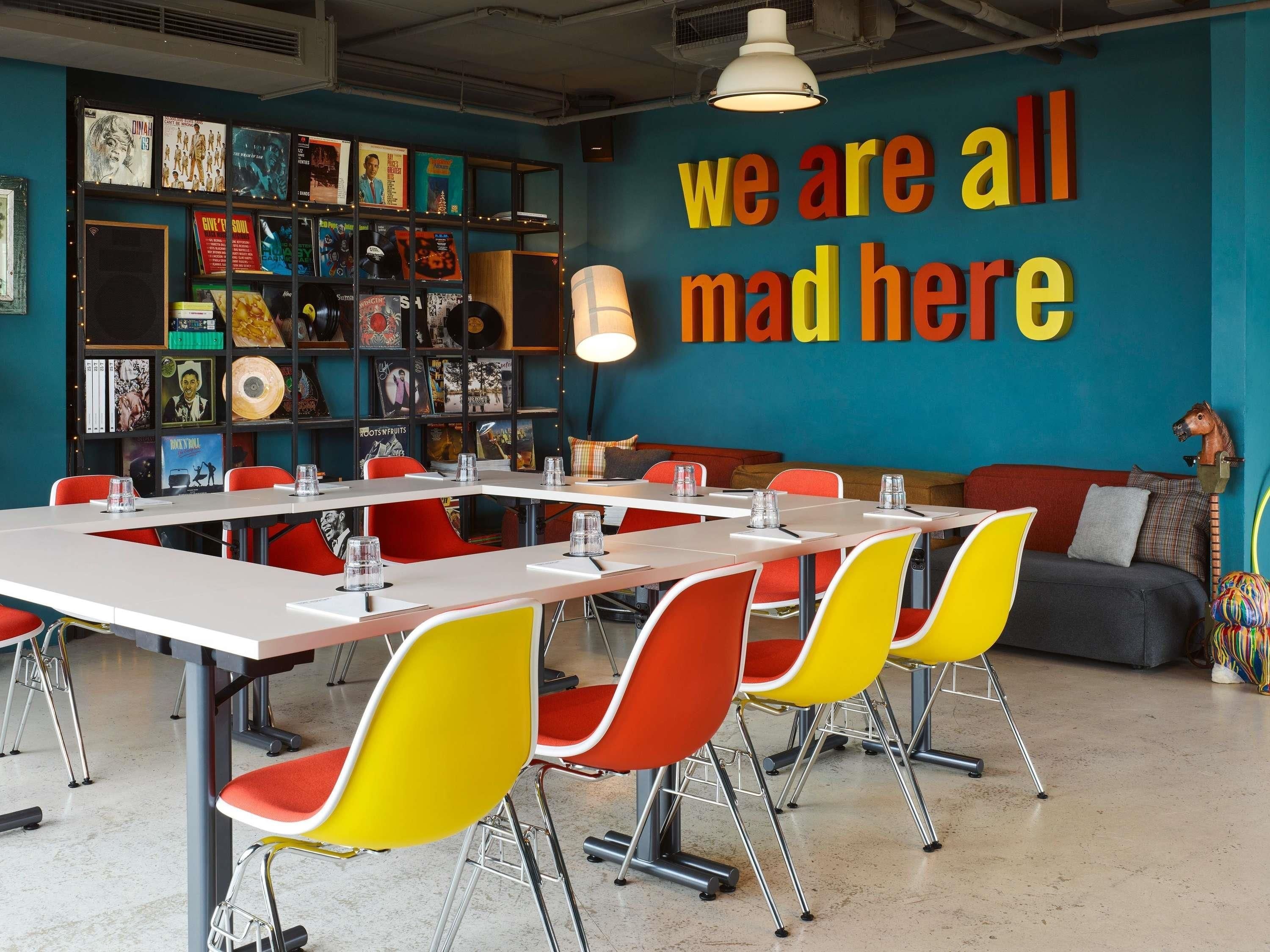 Uma sala de reunião moderna com um grande mural na parede, uma estante com discos de vinil e cadeiras coloridas.
