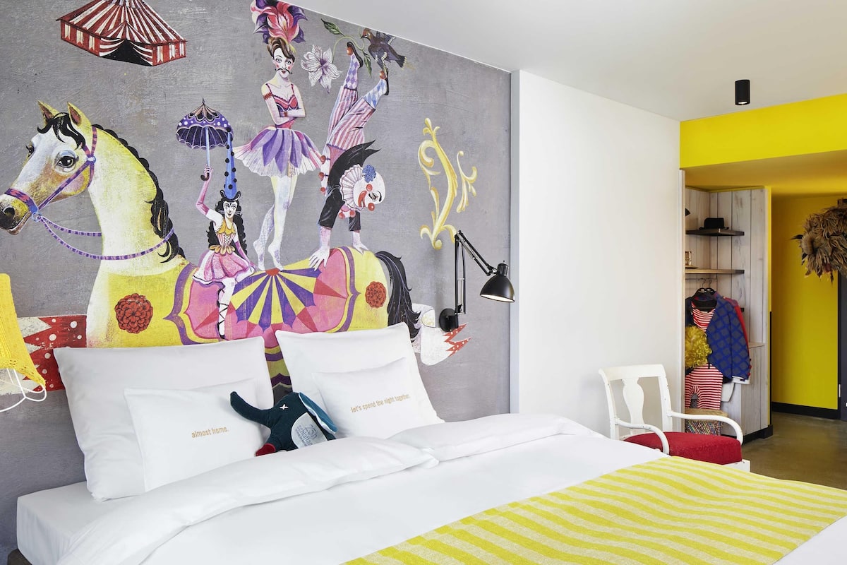 A bed with white bedding, a colorful mural on the wall, and a yellow and white striped rug.