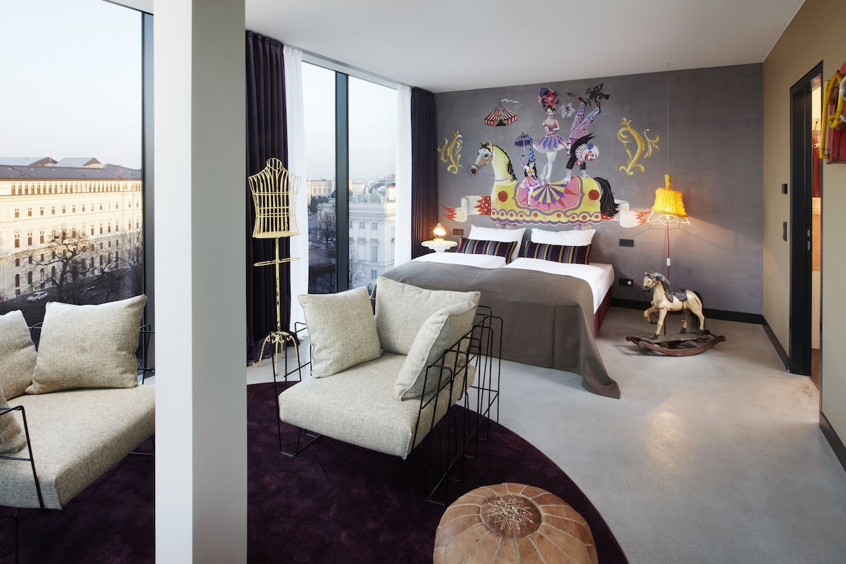 A modern hotel room with a large window, a bed with a canopy, a chair, a small table, and a decorative wall mural.